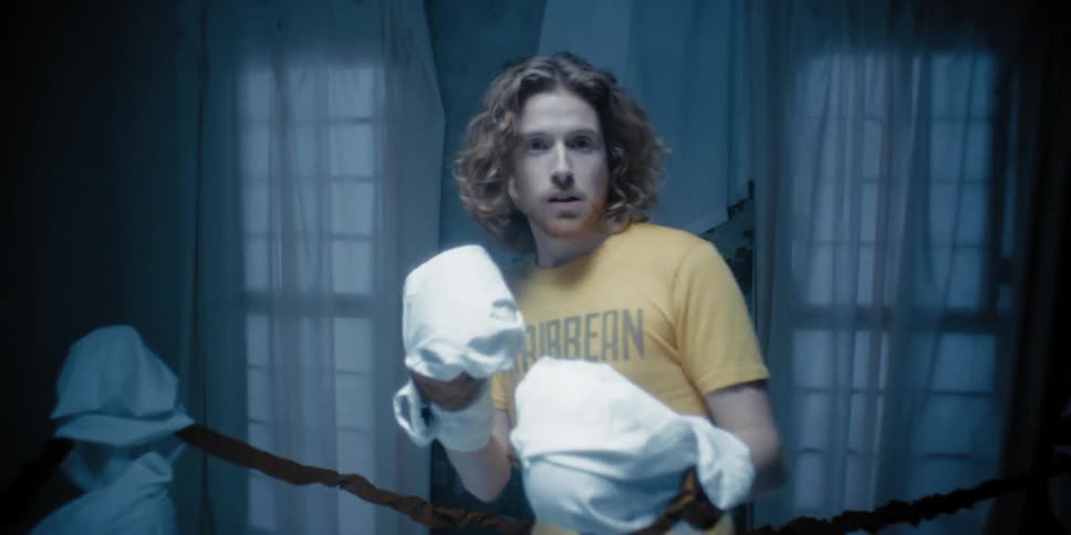 Man in a yellow shirt inside a room resembling a boxing ring wrapping cloth around his hands in preparation for boxing