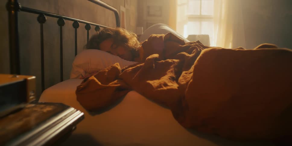 A man peacefully sleeping on his bed covered by a blanket as sunlight streams in through the window