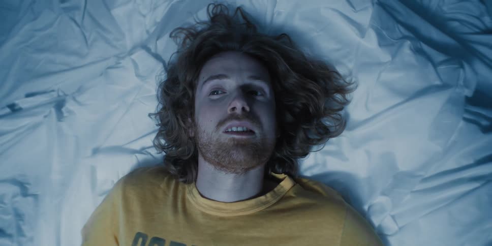 A man slowly opening his eyes and turning his head from the side to look up at the ceiling while flat on his back on top of the bedsheets