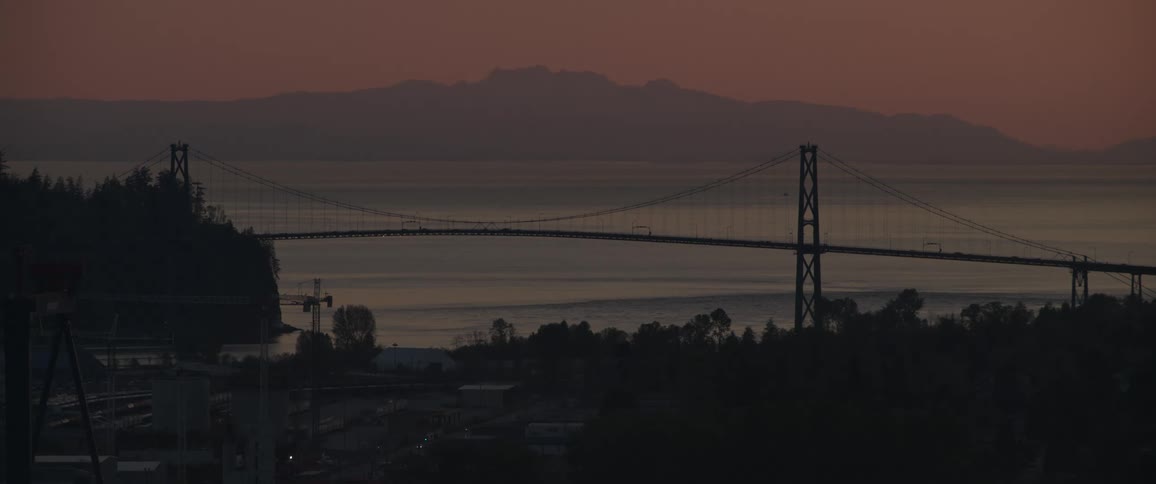 A silhouette view of a tall bridge going over a waterway with a suburb village in the foreground