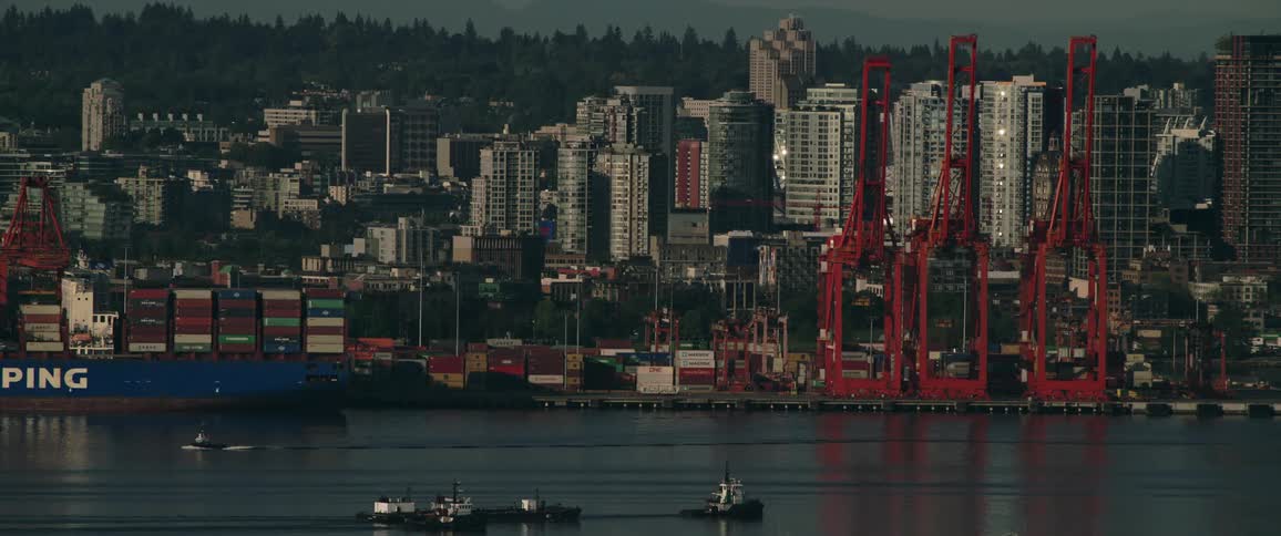 A cargo boat arrives at a city harbor where a larger cargo boat is next to several cranes and is loaded with cargo containers