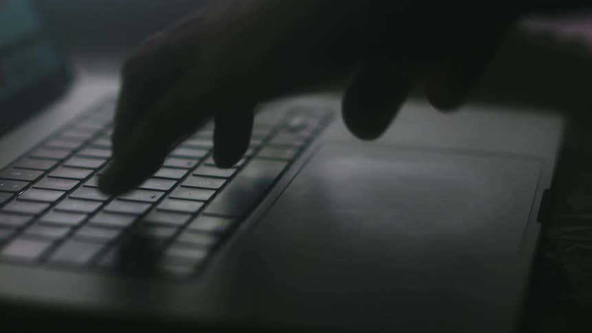 A person uses the keyboard and touchpad of a laptop in a dimly lit room