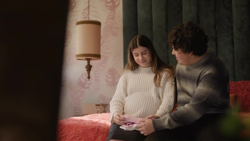 Happy couple seated on the bed the woman being pregnant and they are looking at baby clothes on her lap
