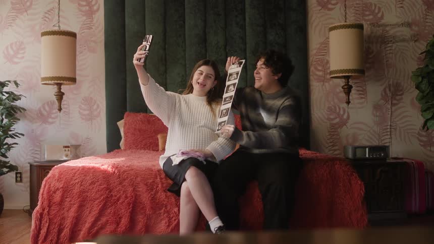 A teenage boy and girl pose together for a selfie while sitting on the edge of a bed