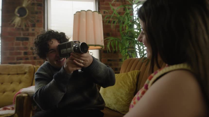 One woman filming another while both laugh and smile sitting on a sofa
