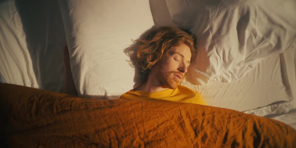 A man sleeping on a bed his head resting and moving on a pillow and covered by a blanket basking in the sunlight