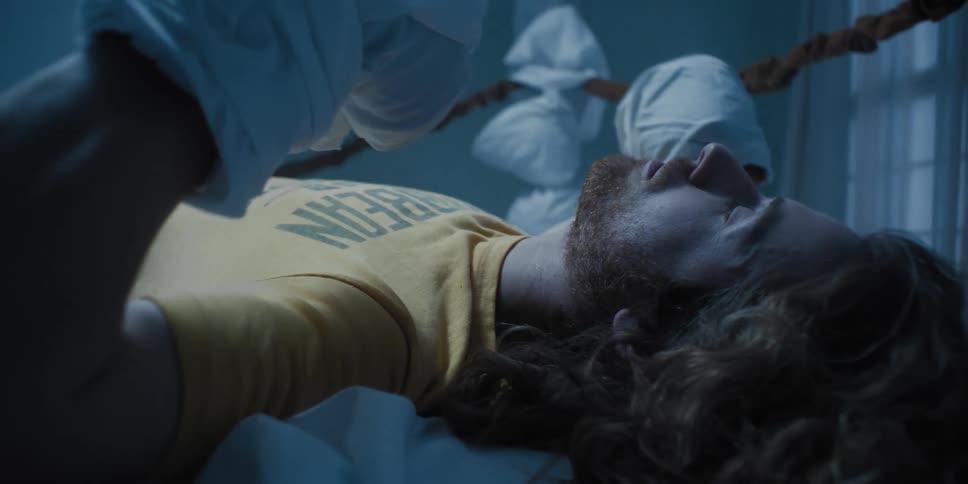 A man with sheets tied around hands like boxing gloves smacks them together while lying on a bed