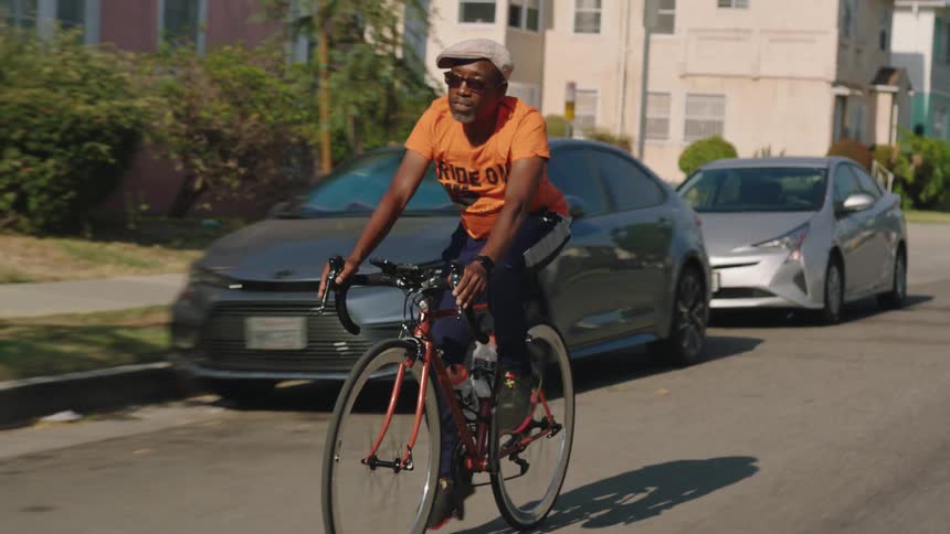 A black man riding his bike through the streets of a sunny residential neighborhood