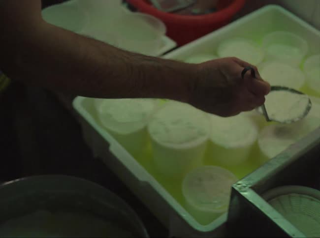 Man scooping tofu from large bucket into smaller containers in a kitchen with a tiled wall