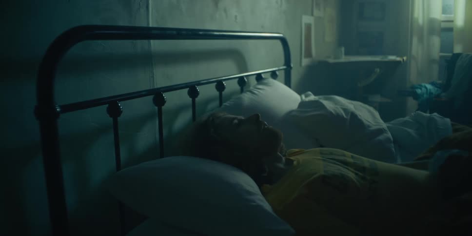 A man flips a pillow over and then lays on the bed in a dark bedroom