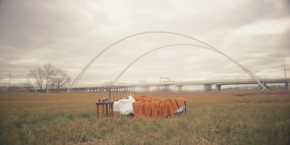 A woman trying to sleep in a bed on a grass field near an arched bridge on a cloudy fall day