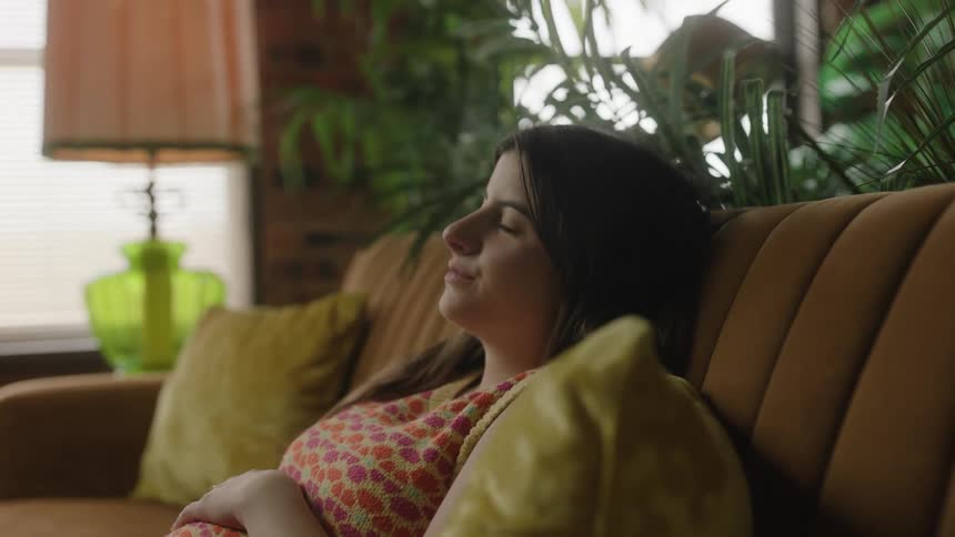 A pregnant woman in slow motion sits on the couch and rubs her belly