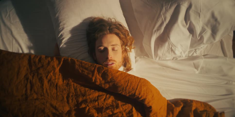 A man asleep in his bed covered by a blanket and illuminated by sunlight