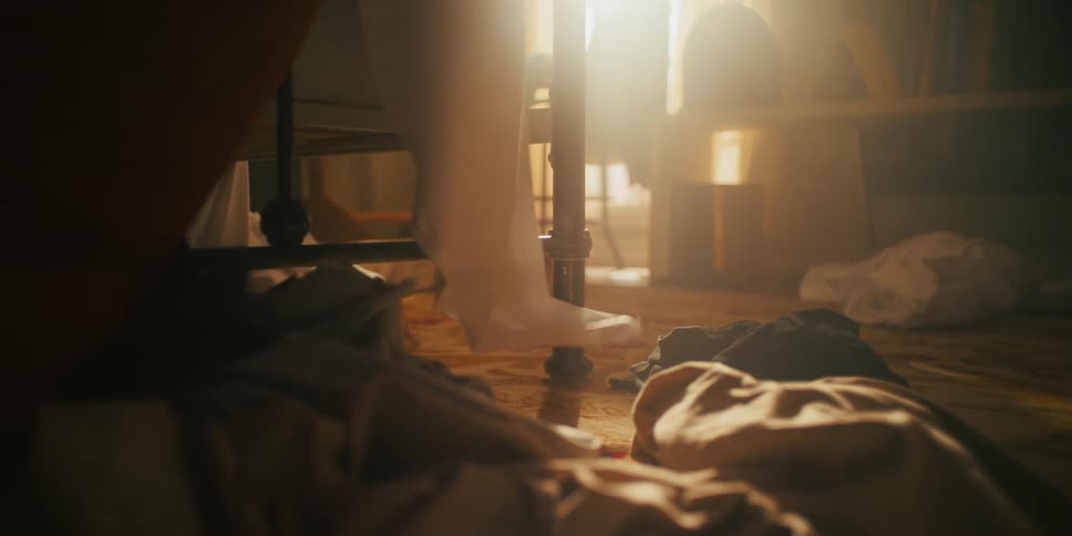 A sheet being pulled up from a metal bed in a room with clothes scattered all over the floor