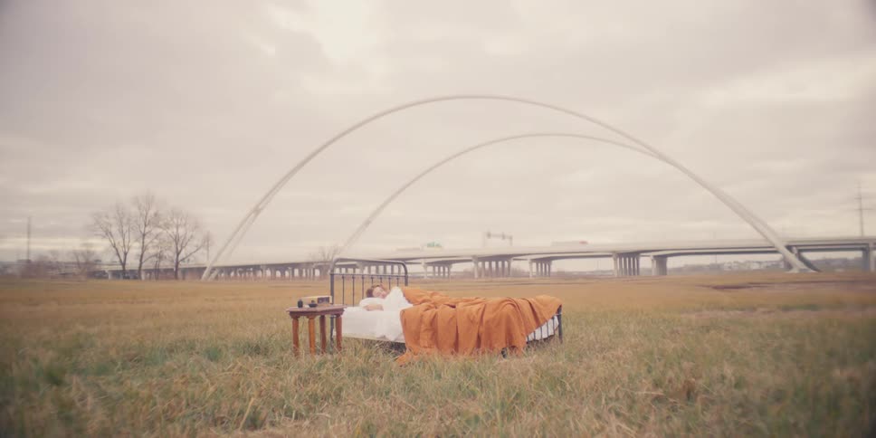 Man sleeping on a bed out on a grass field next to a bridge where cars are passing by