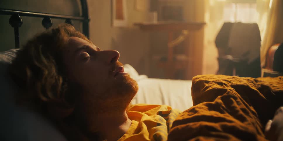 A man lays in bed looking annoyed and not wanting to get up closes his eyes again