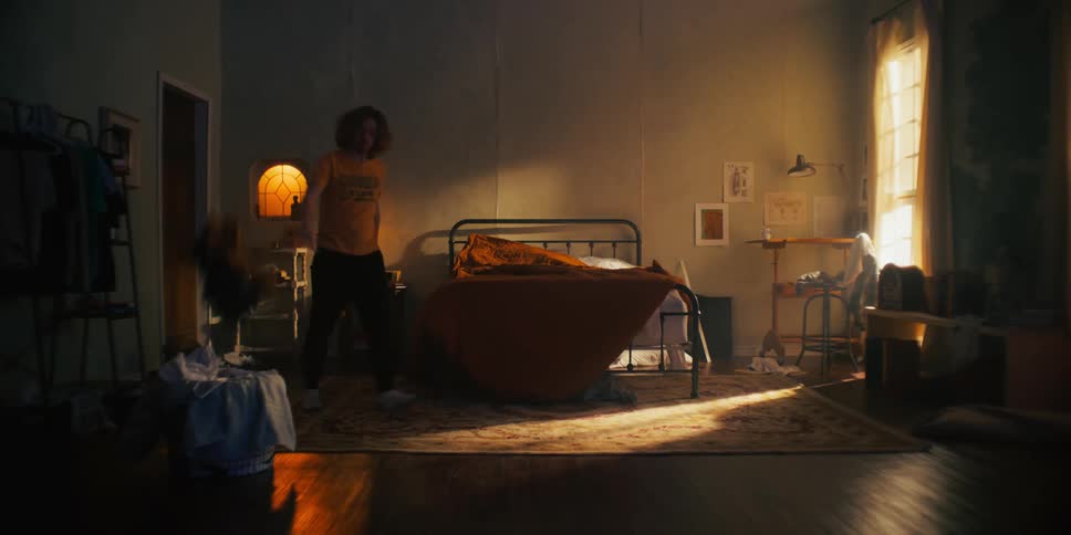 A man is walking around his bedroom cleaning up by throwing things where they go