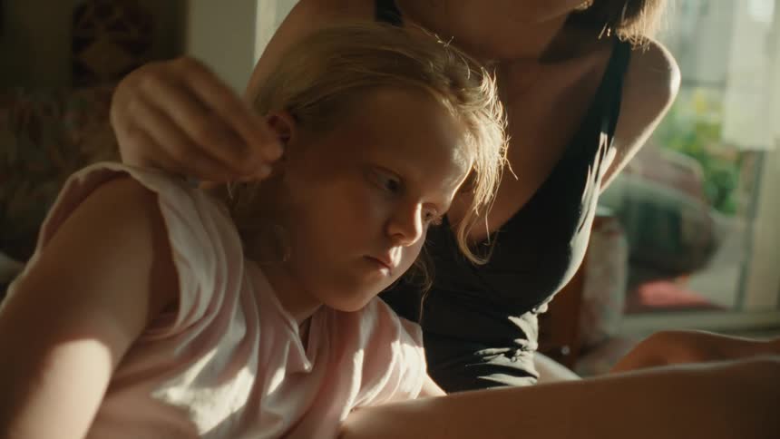 A close up of a young girl and her mother as they sit together while the mother plays with the girls hair