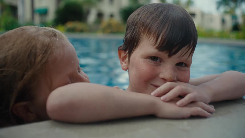 Two kids playing in a pool outdoors with other people in the background