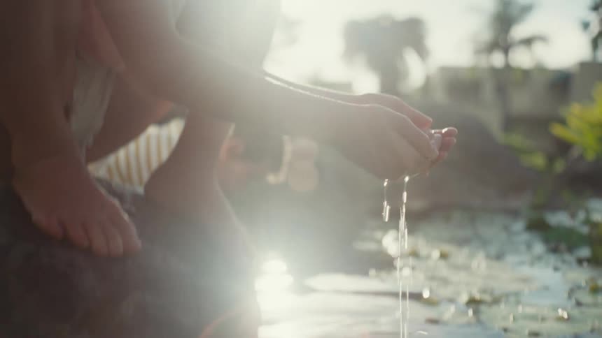 A girl scoops water from a pond with her hands while sitting on a rock during the day