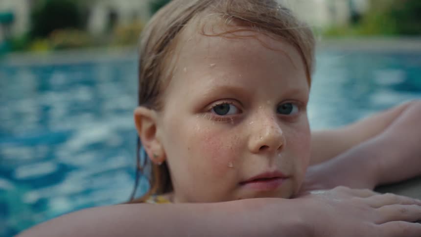 A girl looks into the camera while resting against the side of a swimming pool during the day