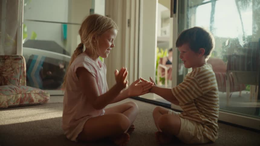 A boy and a girl play a clapping game while sitting on the floor in a house during the day