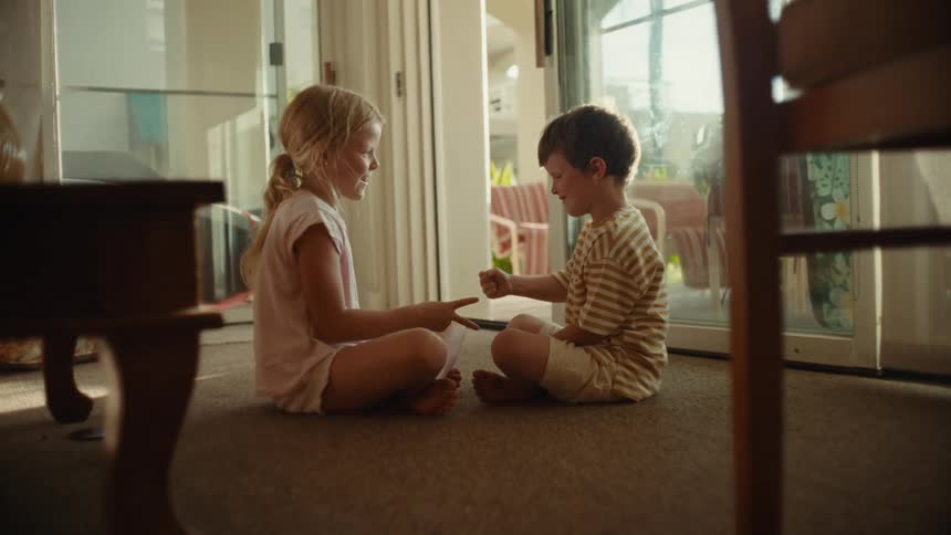 A girl and a boy play a clapping game while sitting on the floor indoors during the day