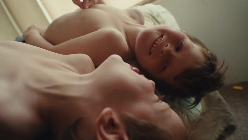 Two shirtless boys hanging their heads off the bed while laughing and smiling