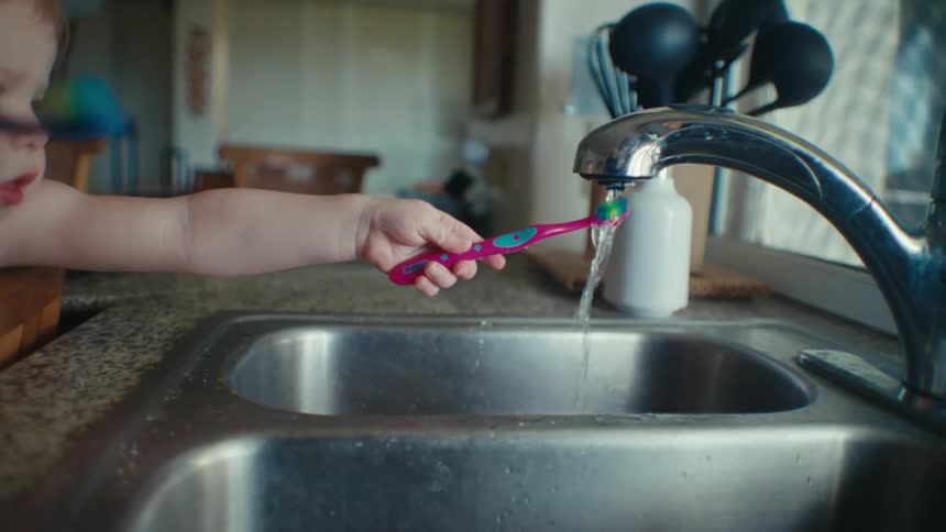 Two young children standing at the kitchen sink reaching to wet their toothbrushes