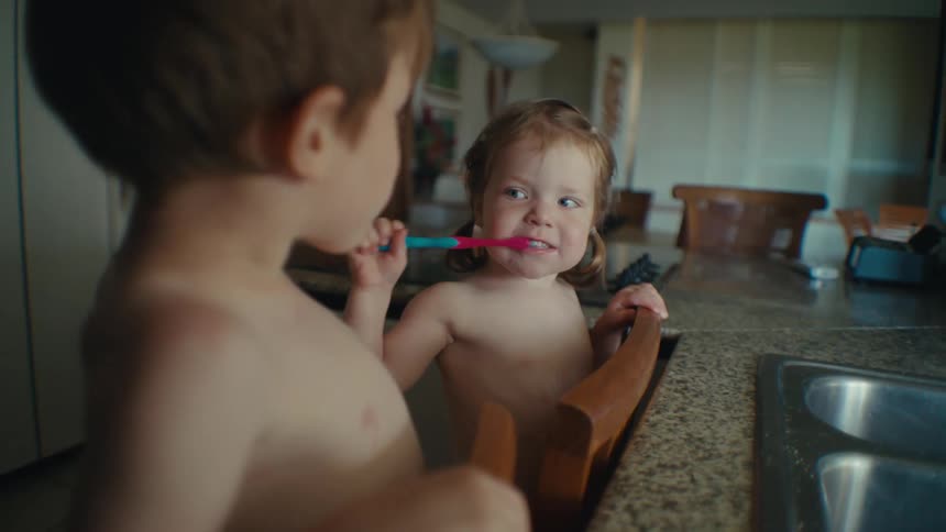 Toddlers brushing teeth together on wooden chairs against the kitchen sink