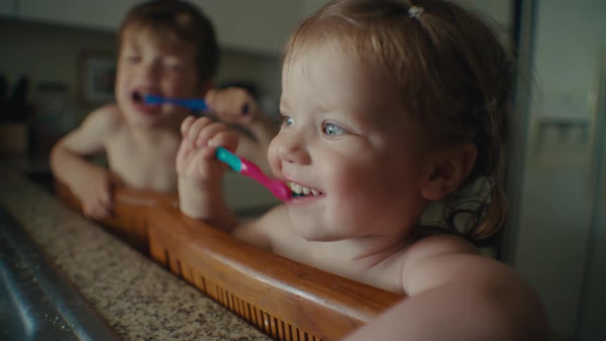 Siblings kneeling on chairs near kitchen sink brush their teeth