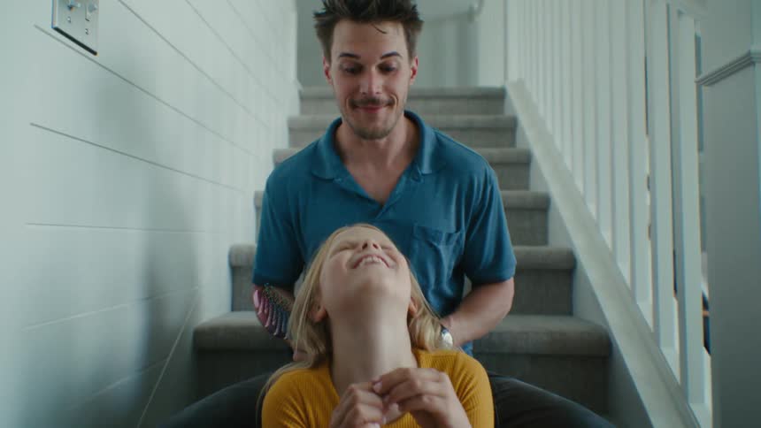 A man brushes the hair of a girl while they sit together on an indoor staircase