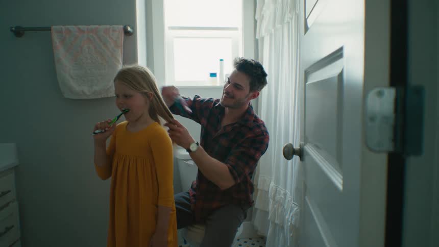 A man brushes the hair of a girl while she brushes her teeth in a home bathroom during the day