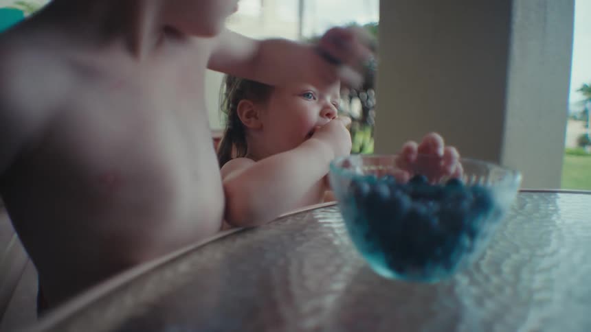 A pair of children eat berries from a glass bowl on a table