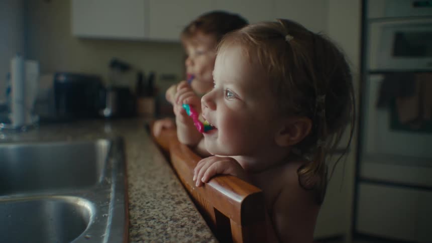 Toddlers standing on a chair brushing their teeth at kitchen sink