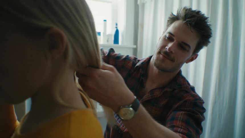 A man brushes the hair of a girl while she brushes her teeth in a bathroom during the day