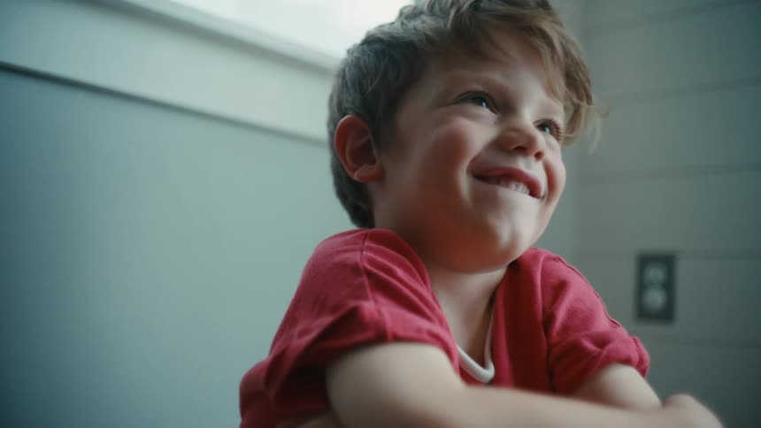 Small boy wearing a red shirt sitting on the floor laughing and gesturing to the camera