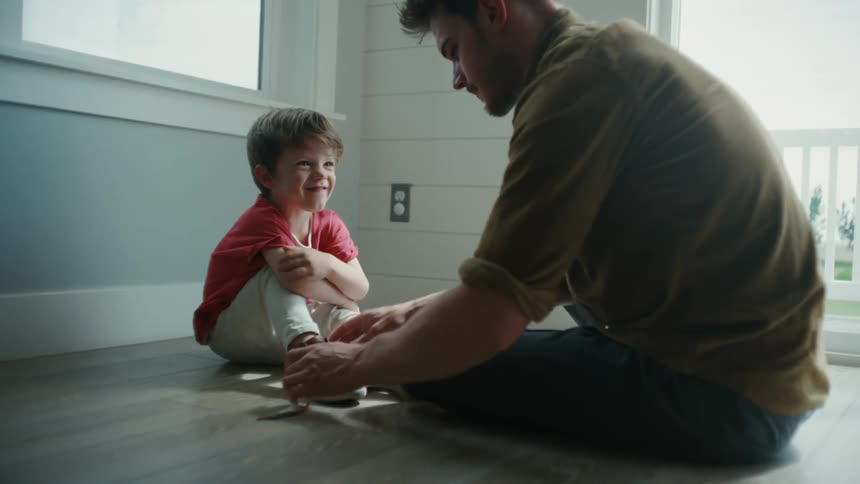 A young Father tying his sons shoe as the child looks up at him and smiles