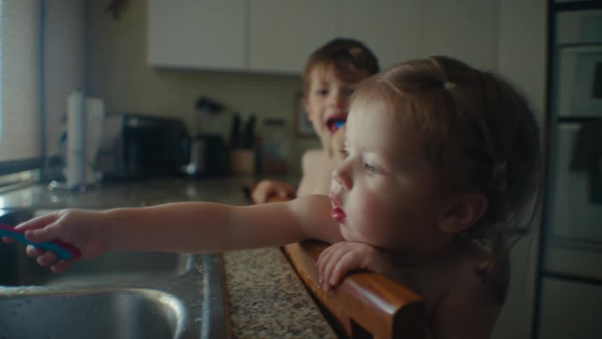 Brother and sister toddlers attempt to brush their teeth at the kitchen sink