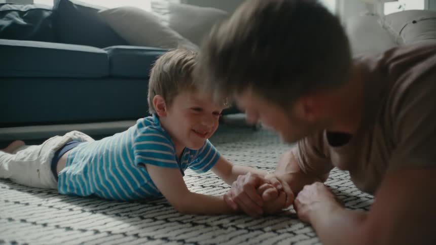A man pretends to struggle while arm wrestling a small child on the carpet in the living room of a house during the day