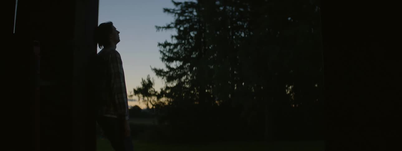 Silhouette of a man outdoors leaning against a wall while smoking a cigarette surrounded by trees