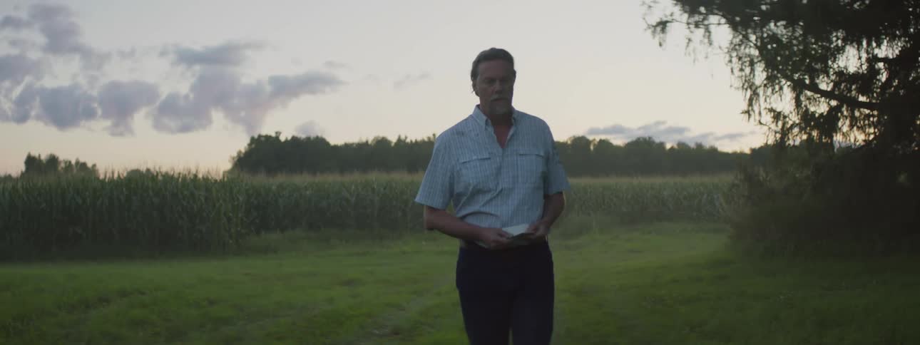 A man stands in a field thinking and tapping a piece of paper in his hand and then turns and walks away