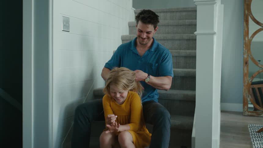 A father and daughter sitting on a staircase in their home while the dad brushes the girls hair and they both smile and laugh