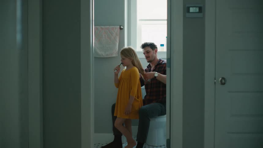 Father brushes his daughters hair while she brushes her teeth in the bathroom