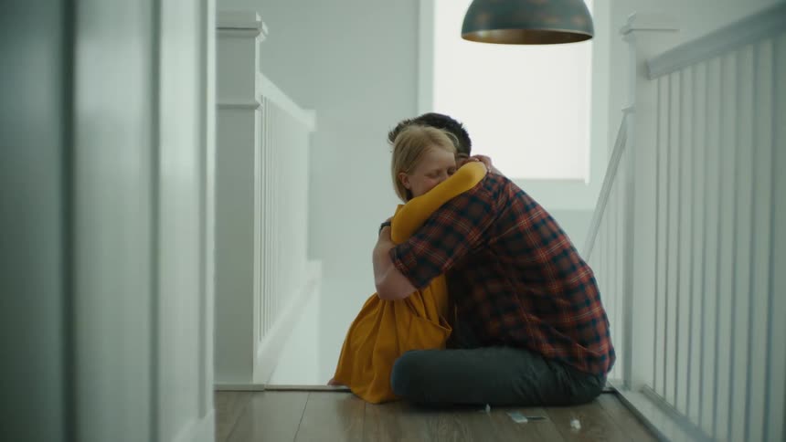 A father kisses his sad daughters hand then hugs her while they are sitting on the floor