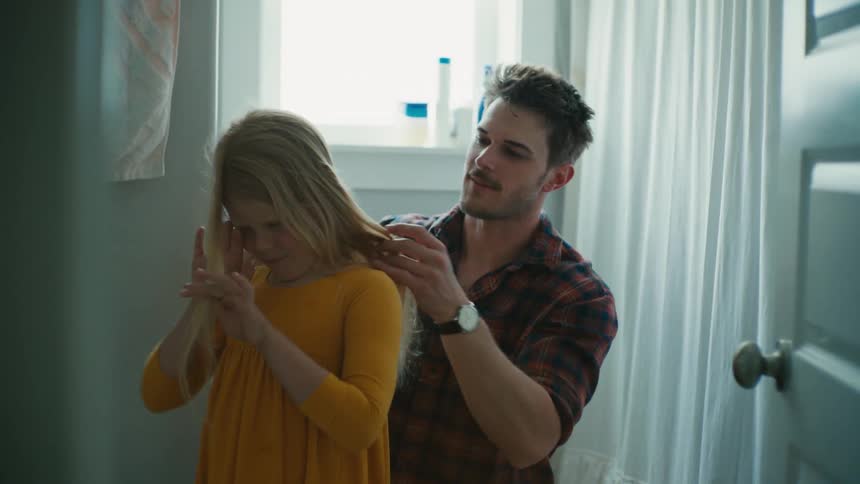 Darkhaired man combing the hair of a blonde girl wearing a yellow dress in a bathroom