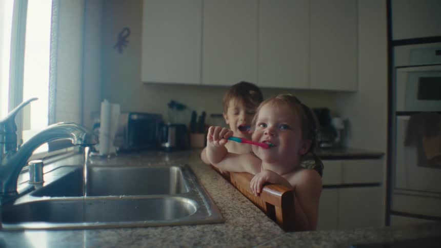 Siblings stand on chair to brush their teeth at the kitchen sink