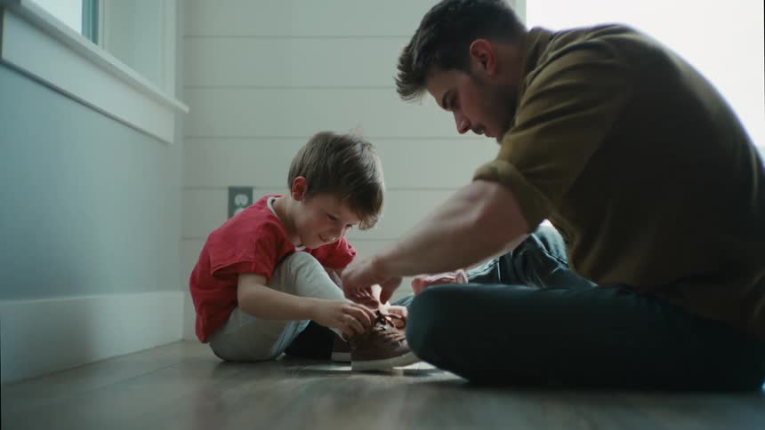 A man helps a young boy to tie his shoes while they sit together on the floor during the day
