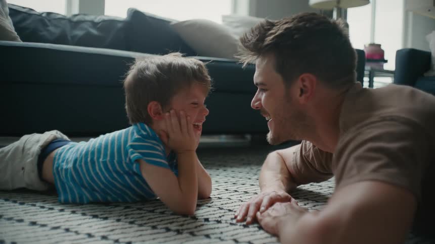 Father and son lie on their stomachs in living room smiling at each other