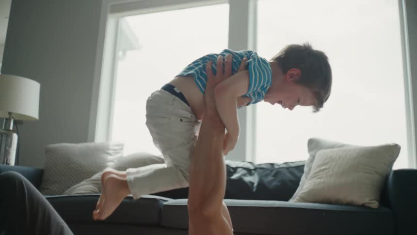 Father lies on his back on floor in living room and lifts son into the air